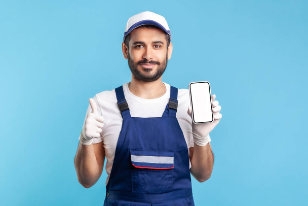 Satisfied consultant in overalls and gloves holding mobile phone with mock up display and showing thumbs up. Good online delivery order app, house repair maintenance services. indoor studio shot - Φωτογραφία, εικόνα