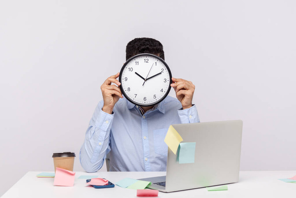 Overtime work. Anonymous man employee sitting in office workplace, hiding face behind clock, sticky notes all around reminding of deadline, stressful job. studio shot isolated on white background - Photo, image