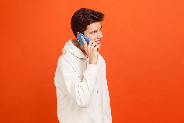 Smiling happy young man in casual style hoodie holding phone near ear, talking with girlfriend, flirting, good connection. Indoor studio shot isolated on orange background - Photo, Image