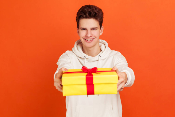 Handsome cheerful teenager in casual sweatshirt holding out yellow gift box with orange ribbon and bow and smiling at camera, congratulation. Indoor studio shot isolated on orange background - Fotografie, Obrázek