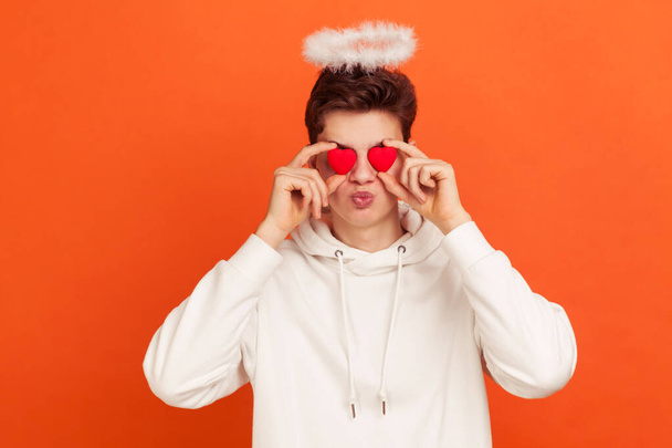 Adorable teenager in casual style hoodie with halo overhead sending kisses holding toy hearts near eyes, man celebrating saint valentine day. Indoor studio shot isolated on orange background - Foto, immagini