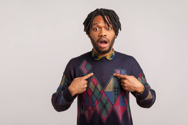 Extremely surprised shocked african man with dreadlocks pointing fingers at himself with widely open eyes and mouth, wondered with choice. Indoor studio shot isolated on gray background - Foto, Bild