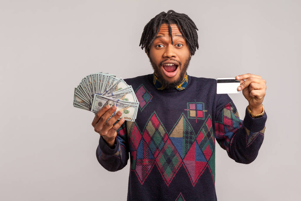 Excited shocked african man with dreadlocks holding in hands batch of dollars and credit card looking at camera, surprised with crediting, deposit. Indoor studio shot isolated on gray background - Zdjęcie, obraz