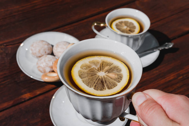 Κρατήστε ένα φλιτζάνι τσάι με λεμόνι στο χέρι σας. Τσάι κόμμα με νόστιμο φαγητό και γλυκά. Κεραμικά λευκό Κύπελλο και σετ τσαγιού. - Φωτογραφία, εικόνα