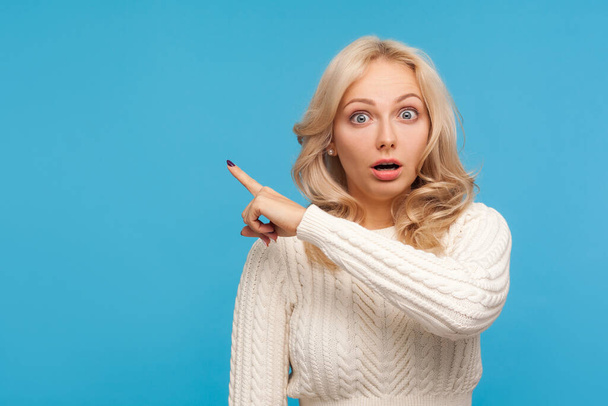 Surprised shocked blond woman in knitted sweater pointing finger away at empty blank for your advertisement, wondered with accessibility. Indoor studio shot isolated on blue background - Photo, image