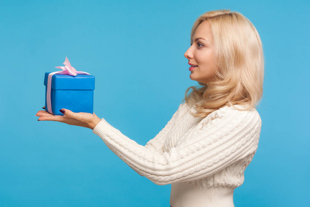 Side view beautiful woman with curly blond hair holding blue gift box in hands, making present, donation, charity concept. Indoor studio shot isolated on blue background - Φωτογραφία, εικόνα