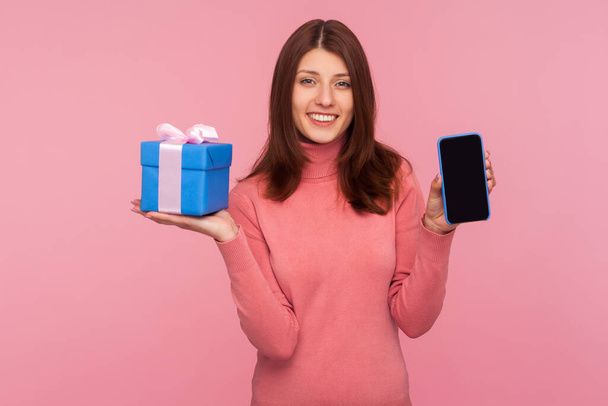 Happy brunette woman in pink sweater holding and showing mobile phone and blue gift box. Indoor studio shot isolated on pink background - Foto, Imagem