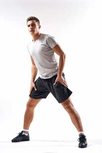 Un hombre deportivo en pantalones cortos y una camiseta larga hace ejercicios sobre un fondo claro - Foto, Imagen