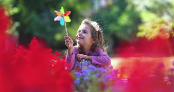Szczęśliwa młoda dziewczyna gra i wieje pinwheel w parku z kolorowymi kwiatami w zwolnionym tempie - Materiał filmowy, wideo