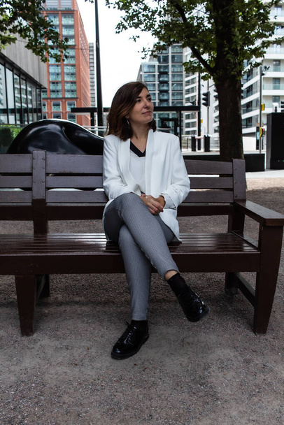 Attractive young female wearing a white suit while sitting on a bench and waiting to start a meeting at her office in London, United Kingdom - Valokuva, kuva