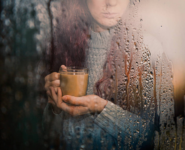 girl with a mug of cocoa looks at the rain through the window - Фото, изображение