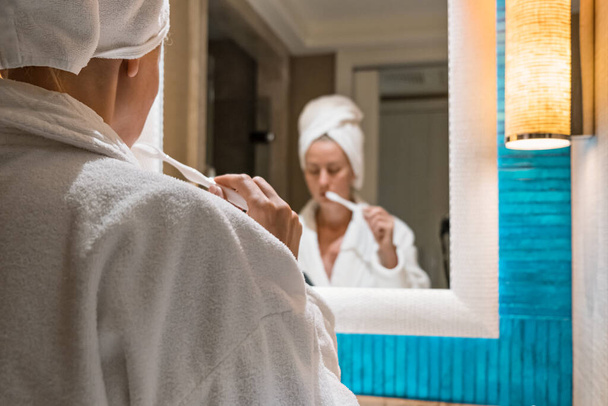 mujer joven en albornoz y toalla cepillándose los dientes delante del espejo - Foto, Imagen