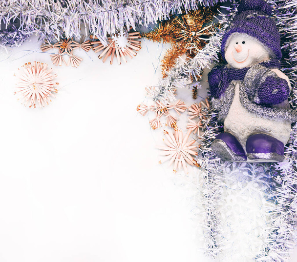Figura de un muñeco de nieve de porcelana en un sombrero azul sobre un fondo blanco. Brillante oropel de Navidad y estrellas artificiales y copos de nieve, juguetes de Navidad. Hermoso fondo de Navidad. - Foto, Imagen