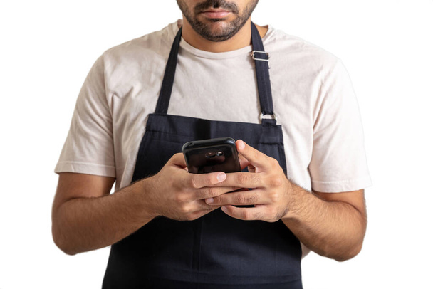 Man holding phone. Body of young waiter with beard and black apron using a mobile isolated on white background. Break time for the worker. Closeup view. - Photo, image