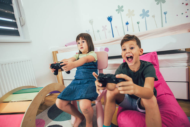 Досить маленька дівчинка і хлопчик грають в ігрову консоль і сміються вдома
 - Фото, зображення