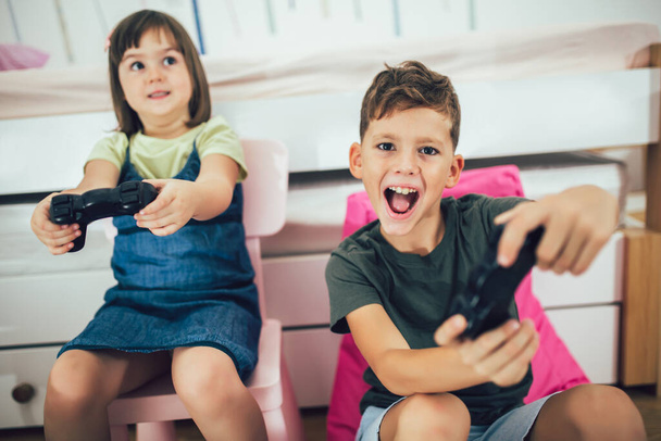 Pretty little girl and boy are playing game console and laughing at home - Фото, изображение