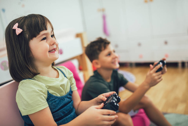 Ładna dziewczynka i chłopiec grają w konsolę do gier i śmieją się w domu - Zdjęcie, obraz