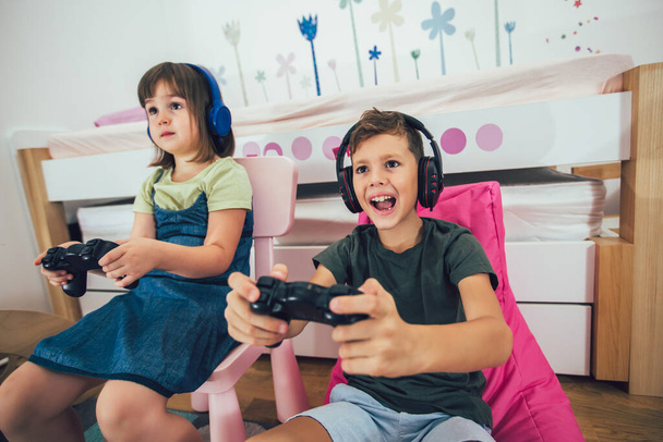 Досить маленька дівчинка і хлопчик грають в ігрову консоль і сміються вдома
 - Фото, зображення