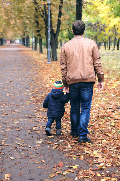 Sonbahar ailesi ormanda yürür. Kuru sarı yaprakları olan güzel bir park. Oğlu ve babası el ele tutuşuyor.. - Fotoğraf, Görsel