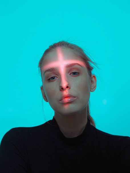 Portrait of young pretty girl with cross shaped red light on her face on blue studio background - Fotoğraf, Görsel