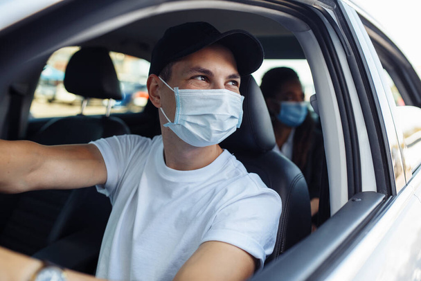 Young taxi driver looks out of a car's window while driving through the city with a passanger wearing a medical mask. Business trips during pandemic, new normal and coronavirus travel safety concept - Foto, Bild
