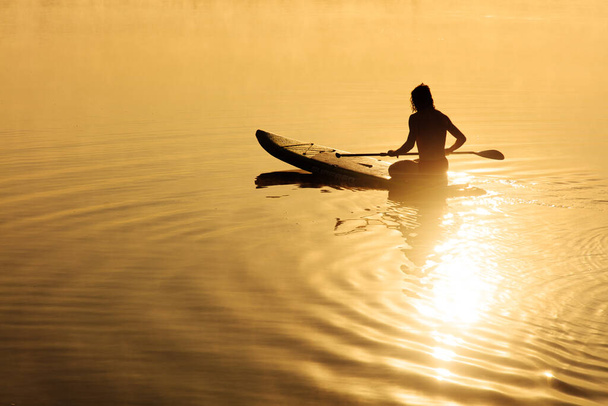 Silueta silného muže sedícího na sup boardu a používajícího dlouhé pádlo k plavání u mlhavého jezera. Úžasný letní východ slunce na pozadí. - Fotografie, Obrázek