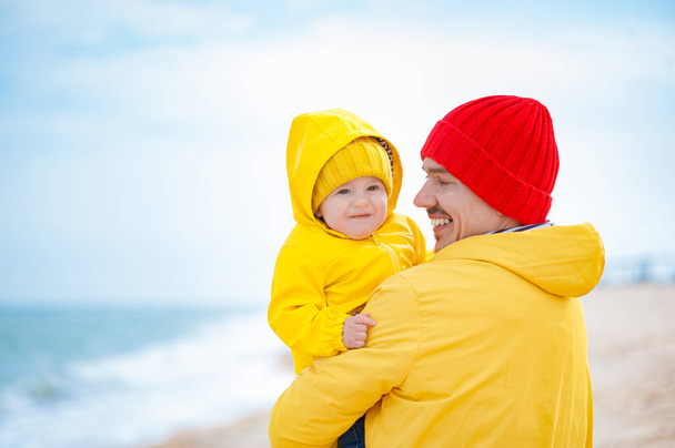 Papa avec un bébé au bord de la mer en vêtements colorés. Journée ensoleillée mais froide et venteuse. - Photo, image