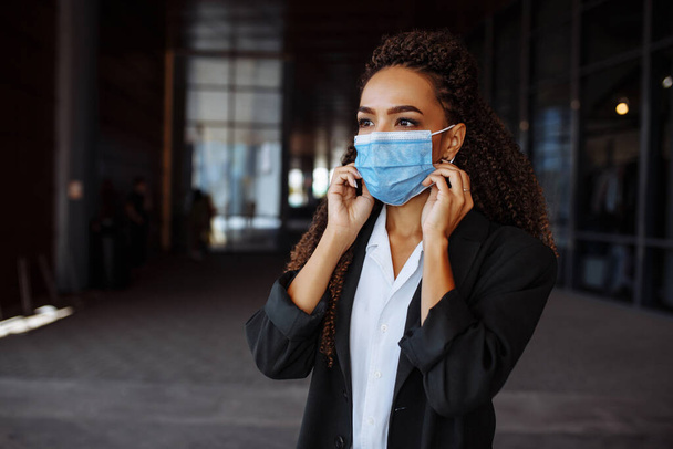 Młoda, oficjalnie wyglądająca kobieta w garniturze dostosowuje maskę medyczną przed biurem, rozmawiając o interesach przez komórkę. Wiodąca praca biznesowa podczas kwarantanny Covid-19 - Zdjęcie, obraz