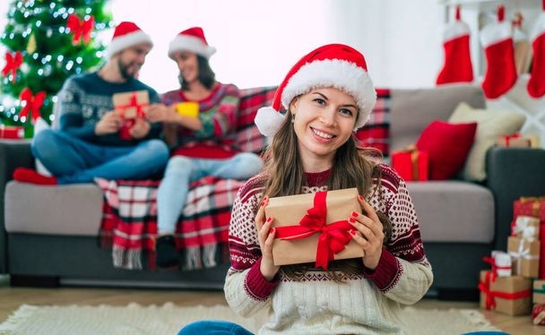 Beautiful young happy smiling woman in a Christmas sweater and Santa hat is holding a colorful gift box in hands at home on the background of her friends and Christmas tree. - Фото, изображение