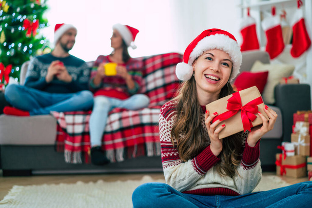Beautiful young happy smiling woman in a Christmas sweater and Santa hat is holding a colorful gift box in hands at home on the background of her friends and Christmas tree. - Photo, Image