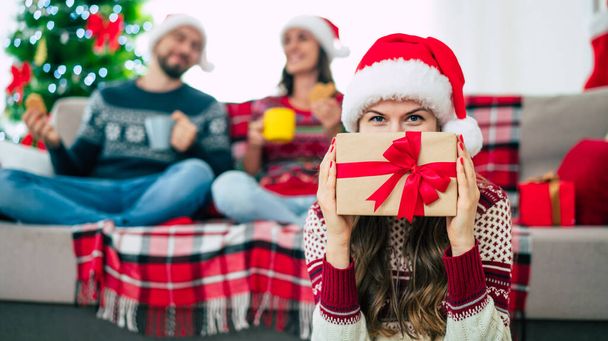 Close up photo of beautiful young happy smiling woman in a Christmas sweater and Santa hat is holding a gift box in hands at home on the background of her friends and Christmas tree. - 写真・画像