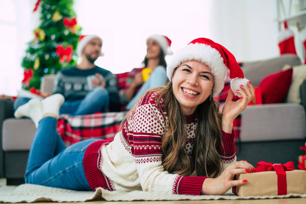 Beautiful young happy smiling woman in a Christmas sweater and Santa hat is holding a colorful gift box in hands at home on the background of her friends and Christmas tree. - Zdjęcie, obraz