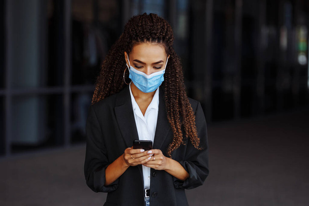 Zbliżenie młodej bizneswoman smsującej telefonem komórkowym w masce medycznej. Piękna dziewczyna rozmawiająca na smartfonie przed biurem. Koncepcja biznesowa Covid-19 - Zdjęcie, obraz