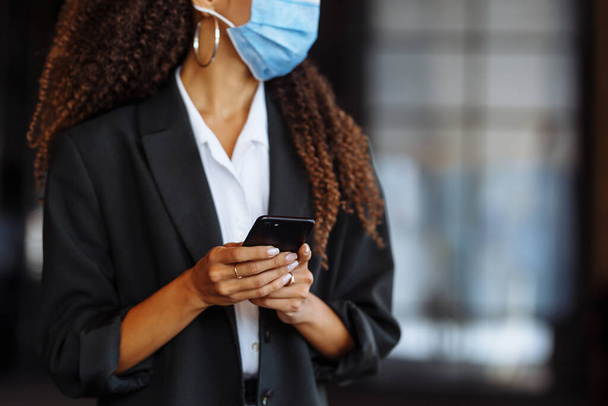 Zbliżenie młodej bizneswoman smsującej telefonem komórkowym w masce medycznej. Piękna dziewczyna rozmawiająca na smartfonie przed biurem. Koncepcja biznesowa Covid-19 - Zdjęcie, obraz