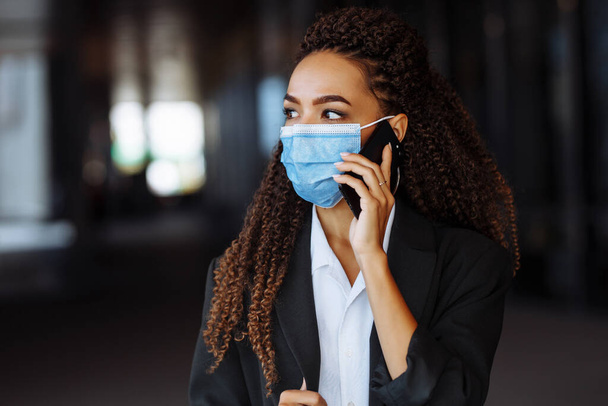 Ritratto di una giovane donna attraente che parla di affari sul cellulare indossando una maschera medica sterile fuori dall'edificio degli uffici. Principali attività durante il concetto di pandemia di Covid-19 - Foto, immagini