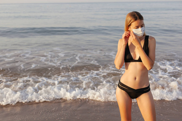 Beautiful fit girl standing by the sea and putting on medical mask. New rules of traveling 2020. Coronavirus concept. - Foto, Bild