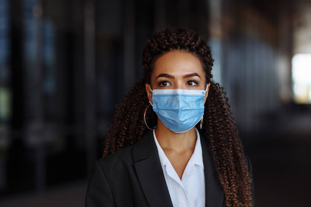 Retrato de una joven mujer de negocios con una máscara médica estéril parada fuera del edificio de oficinas. Prevención de la propagación del coronavirus durante la cuarentena pandémica mundial. Empresa líder Covid-19 - Foto, imagen