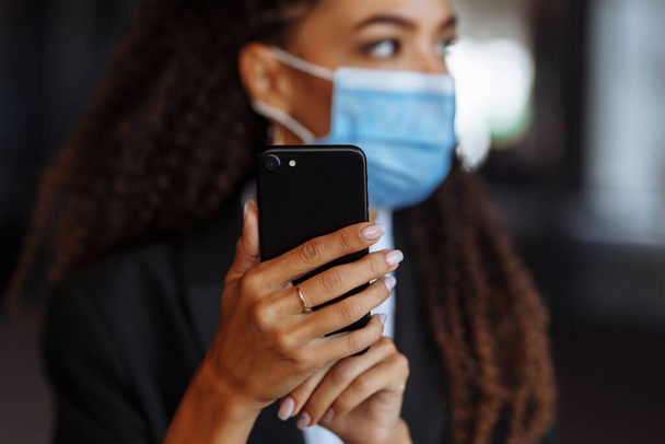 Młoda bizneswoman w masce medycznej stoi w pobliżu centrum biurowego. Oficjalnie wyglądająca dziewczyna z telefonem w rękach czeka na zewnątrz. Wiodący biznes podczas pandemii Covid-19 - Zdjęcie, obraz