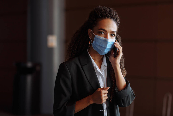 Portrait of a young attractive woman talking business on the mobile phone wearing a medical sterile mask outside the office building. Leading business during Covid-19 pandemic concept - Φωτογραφία, εικόνα