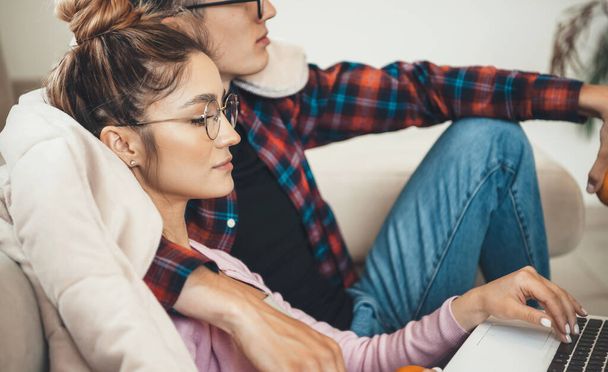 Side view photo of a caucasian woman with glasses embraced by her lover sitting on the couch and using a laptop - Fotografie, Obrázek
