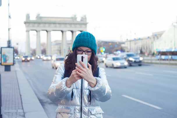 Дівчина в пуховику і капелюх гуляє по місту і фотографується на смартфоні
, - Фото, зображення