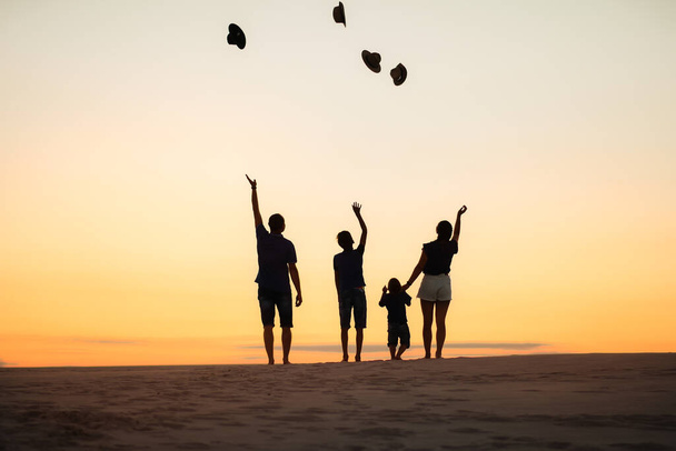 Silhouette of a family at sunset on the Sands. Mom and dad and two sons throw up their hats. - Photo, Image