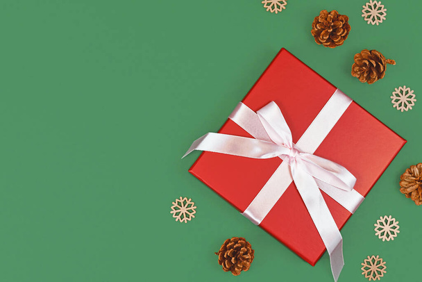 Red Christmas gift box with ribbon surrounded by seasonal pine cones and wooden snowflake ornaments on side of green background with empty copy space - Foto, Imagem
