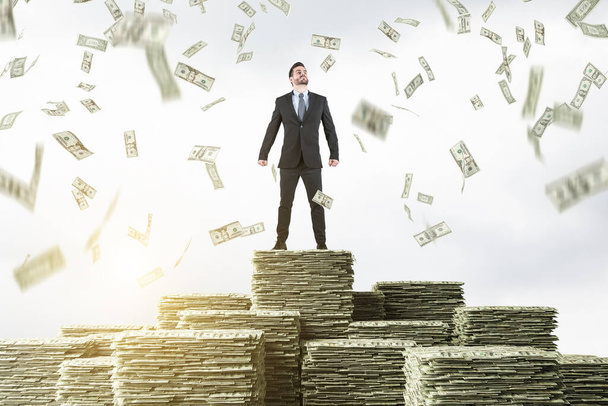 Confident young businessman standing on stacks of money under dollar rain. Concept of investment and success. Toned image - Фото, зображення