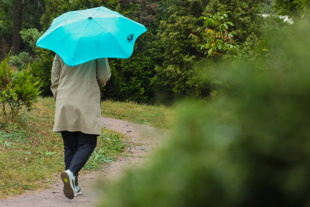 Μια γυναίκα με μπεζ παλτό περπατάει στον κήπο του φθινοπώρου κάτω από μια πράσινη ομπρέλα μια βροχερή μέρα.. - Φωτογραφία, εικόνα
