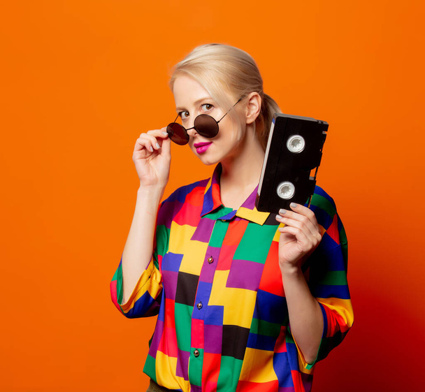 Style blonde in 90s clothes with VHS cassette on orange background - Fotografie, Obrázek