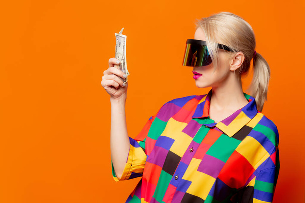 Style blonde in 90s clothes and VR glasses with money on orange background - Photo, Image