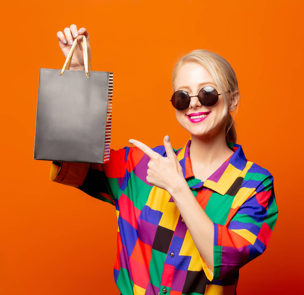 Style blonde in 90s shirt and sunglasses with shopping bag on orange background - Photo, Image