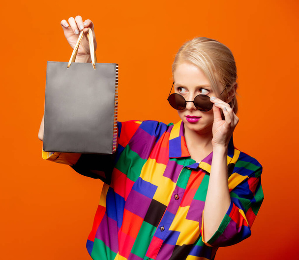 Στυλ ξανθιά σε μπλούζα της δεκαετίας του '90 και γυαλιά ηλίου με τσάντα για ψώνια σε πορτοκαλί φόντο - Φωτογραφία, εικόνα