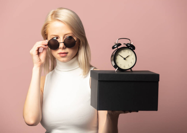 Style blonde in top and sunglasses with alarm clock and black gift box on pink background - Foto, afbeelding